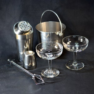 Op de afbeelding: Een zilveren cocktailshaker, een ijs emmer en twee martini glazen op een zwart oppervlak. De shaker heeft een zeefdeksel en een zilveren cocktail muddler.
