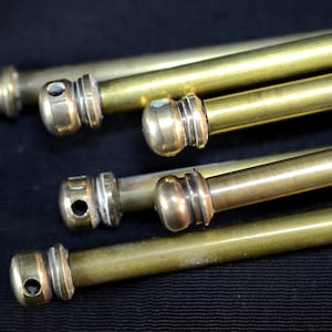 May include: Close-up of several brass rods with decorative ends. The rods are cylindrical with a smooth, polished finish. Each rod has a rounded end with a small hole and a decorative collar. The rods are arranged diagonally against a dark background.