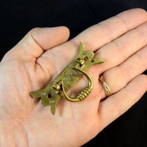 Può includere: Una maniglia per cassetto in ottone con un design decorativo e un'impugnatura ad anello. La maniglia ha la forma di un uccello stilizzato con un corpo lungo e stretto e una coda appuntita.