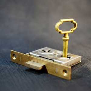 May include: A brass key is inserted into a brass door lock. The key has a decorative, ornate design. The lock is attached to a brass plate.