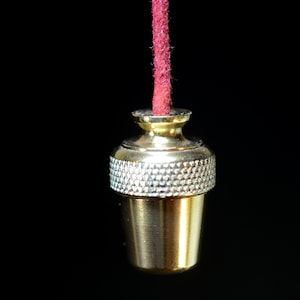 Może przedstawiać: Złoty ściągacz do światła z teksturowanym srebrnym paskiem i czerwonym sznurkiem. Ściągacz ma klasyczny design, z cylindryczną podstawą i ozdobną górą. Tło jest czarne.