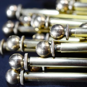 Könnte beinhalten: Nahaufnahme mehrerer Gardinenstangen-Endstücke aus Messing mit kugelförmigem Design. Die Stangen sind zylindrisch mit einem Gewindeende und einem dekorativen, geriffelten Kragen. Die Endstücke sind glänzend, metallisch goldfarben, und der Hintergrund ist dunkelblau.