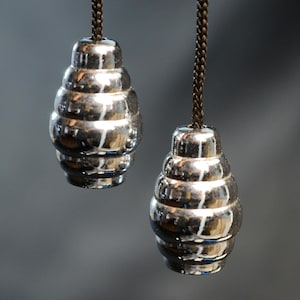 Puede incluir: Dos tiradores de luz plateados, con un diseño en capas, colgando sobre un fondo gris. Los tiradores tienen forma de panal y están unidos a cuerdas marrones. La superficie reflectante crea un efecto brillante. La imagen es un primer plano.