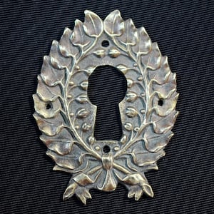Op de afbeelding: Ovale antieke sleutelgatplaat in bronskleur. De plaat heeft een gedetailleerd ontwerp van bladeren en een strik. Het sleutelgat bevindt zich in het midden, met kleine gaten voor bevestiging. De afwerking heeft een vintage uitstraling.