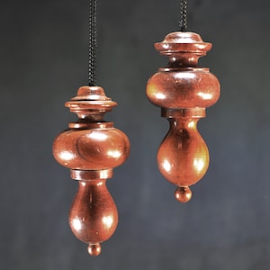Puede incluir: Dos adornos de péndulo de madera colgando de cordones negros. Los adornos son marrones y tienen una forma bulbosa con una parte superior decorativa.