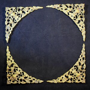 May include: Four decorative, gold-coloured corner brackets arranged to form a square. Each bracket displays detailed floral and scrollwork patterns. The brackets are set against a dark background, accentuating their intricate design.