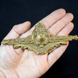 Könnte beinhalten: Verziertes Möbelbeschlagteil aus Messing mit einem ovalen Mittelteil und dekorativen Verzierungen. Das Teil wird in einer Hand vor schwarzem Hintergrund gehalten. Das Teil ist etwa 15 cm lang.