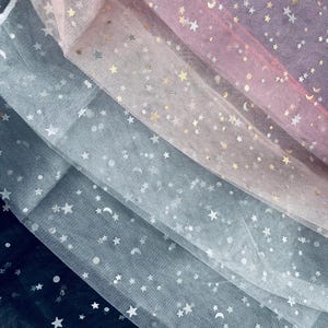 May include: Close-up of layered tulle fabric in shades of pink, white, gray, and navy blue. The tulle is decorated with metallic gold stars and crescent moons, creating a celestial pattern. The fabric is soft and flowing.