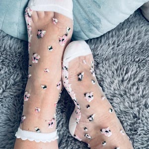 Pode incluir: Meias transparentes com punhos brancos e um delicado padrão floral em rosa e preto. As meias têm uma borda ondulada. O fundo inclui uma almofada azul e uma superfície cinza texturizada.
