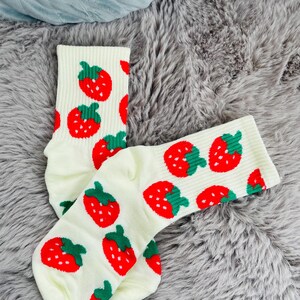 May include: A pair of cream-colored socks with a repeating pattern of red strawberries with green stems and leaves. The socks are displayed on a gray, fluffy surface.