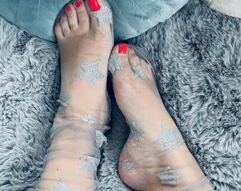 Può includere: Calzini trasparenti grigio chiaro adornati da disegni di stelle glitterate argentate. I calzini sono indossati su piedi con smalto rosso. Lo sfondo comprende un tappeto grigio e soffice e un cuscino verde acqua.