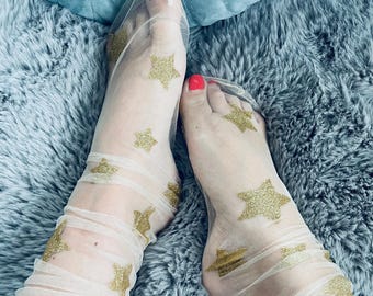 Può includere: Calzini trasparenti beige chiaro con motivi a stella dorati glitterati. I calzini sono arricciati intorno alle caviglie. L'immagine mostra un primo piano dei piedi e delle gambe. Le unghie dei piedi sono dipinte di rosso.