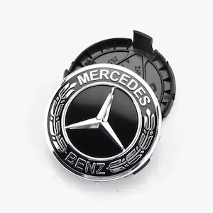 Könnte beinhalten: Eine schwarze und silberne Mercedes-Benz Radnabenabdeckung. Die Kappe zeigt das Mercedes-Benz-Logo, einen dreizackigen Stern in einem Kreis, sowie die Wörter "MERCEDES" und "BENZ". Die Kappe ist vor einem weißen Hintergrund.