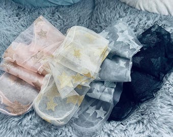 Può includere: Assortimento di rotoli di tessuto trasparente in vari colori, tra cui rosa, bianco, grigio e nero. Ogni rotolo è decorato con motivi a stella glitterati. I tessuti sono disposti su una superficie morbida e strutturata.