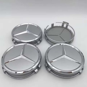 May include: Four silver-colored Mercedes-Benz wheel center caps. Each cap features a three-pointed star emblem in the center, surrounded by a polished chrome ring. One cap is disassembled, showing the back.