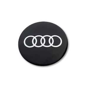 Pode incluir: Um emblema circular preto com o logótipo da Audi em branco, composto por quatro anéis entrelaçados centrados na superfície. O design é plano e minimalista, com um acabamento liso e brilhante que realça o contraste entre o fundo preto e os anéis brancos.