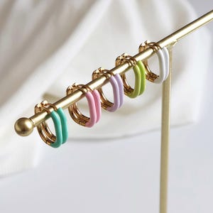 Puede incluir: Exhibición de seis pendientes de aro dorados con detalles de esmalte de colores. Los pendientes están colocados en un expositor dorado. Los colores del esmalte incluyen turquesa, rosa, lavanda, verde lima y blanco.