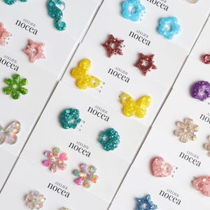 Peut inclure: Une collection d'embellissements décoratifs de différentes formes, dont des hippocampes, des étoiles, des papillons et des fleurs. Les embellissements sont dans une variété de couleurs, dont le teal scintillant, le rose, le jaune et le violet. La marque "nocea" est imprimée sur les cartes.