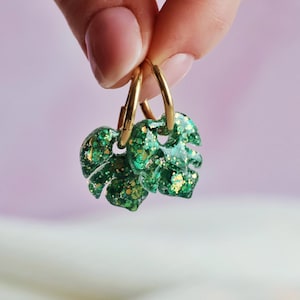 Può includere: Un paio di orecchini a cerchio con ciondoli a forma di foglie di monstera. I ciondoli a foglia sono realizzati in resina verde traslucida con scaglie di glitter dorato, sospesi a semplici cerchi in metallo color oro.