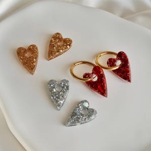 Op de afbeelding: Een verzameling hartvormige oorbellen en bedels in goud, zilver en rode glitter. De oorbellen hebben gouden hoepels. De items worden gepresenteerd op een wit, gebogen oppervlak.