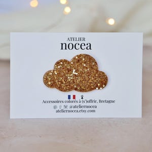 以下が含まれることがあります： 白いカードに金色のグリッター雲型のアクセサリー。カードには「ATELIER nocea」の文字とフランス語の追加テキストが記載されています。アクセサリーはおそらくブローチまたはヘアクリップです。背景はぼやけており、ボケライトがあります。