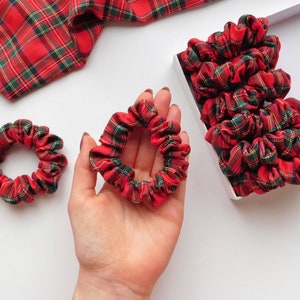 May include: Red, green, and yellow plaid scrunchies. One scrunchie is held in a hand, two are on a white surface, and a box of scrunchies is in the background. A piece of plaid fabric is in the upper left corner.
