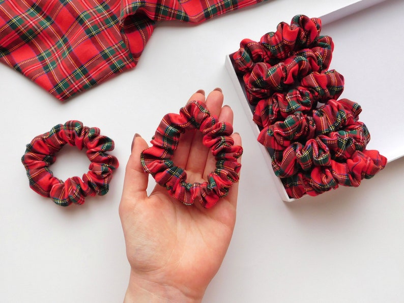 May include: Red, green, and yellow plaid scrunchies. One scrunchie is held in a hand, two are on a white surface, and a box of scrunchies is in the background. A piece of plaid fabric is in the upper left corner.