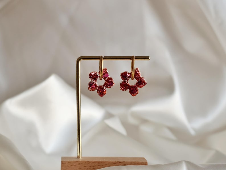 Peut inclure: Une paire de boucles d'oreilles créoles dorées avec des pendentifs en forme de fleur. Chaque pendentif est incrusté de petits cristaux rouges étincelants disposés en une fleur à cinq pétales. Présentées sur un support en métal doré.