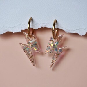 May include: A pair of gold-toned hoop earrings featuring lightning bolt-shaped pendants. The pendants are made of a clear, resin-like material embedded with iridescent, multi-colored glitter flakes that catch the light with various sparkles.