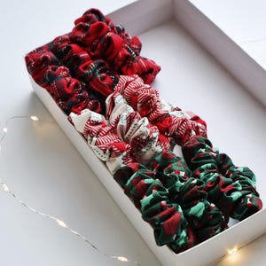 May include: A collection of fabric scrunchies in a white rectangular box. The scrunchies feature various patterns in red, white, and green, including plaid and festive designs. A string of fairy lights is visible in the background.