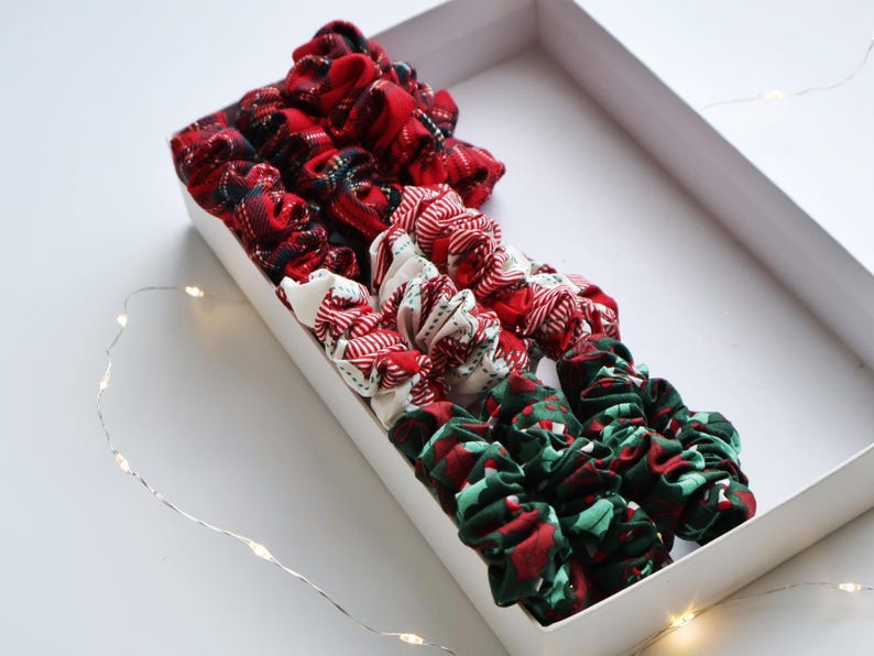 May include: A collection of fabric scrunchies in a white rectangular box. The scrunchies feature various patterns in red, white, and green, including plaid and festive designs. A string of fairy lights is visible in the background.