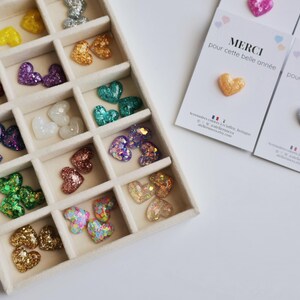 May include: A variety of heart-shaped embellishments in a divided tray. The hearts are in various colors, including gold, silver, pink, purple, and green. Some hearts are glittery. Cards with the text "MERCI pour cette belle ann&eacute;e" are also present.