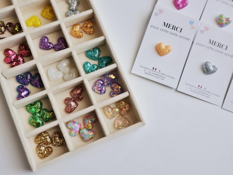 May include: A variety of heart-shaped embellishments in a divided tray. The hearts are in various colors, including gold, silver, pink, purple, and green. Some hearts are glittery. Cards with the text "MERCI pour cette belle ann&eacute;e" are also present.