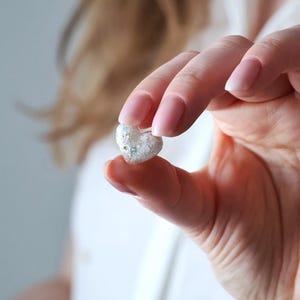 May include: A small, heart-shaped object with a clear, glittery appearance. The object is held between fingers, showcasing its delicate details and sparkling texture. The heart is filled with small, colorful specks, adding to its charm.