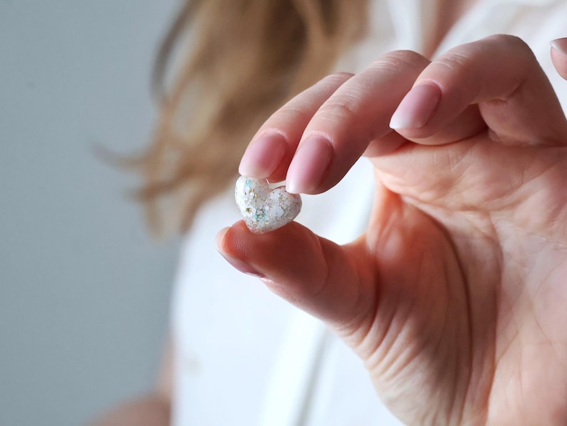 May include: A small, heart-shaped object with a clear, glittery appearance. The object is held between fingers, showcasing its delicate details and sparkling texture. The heart is filled with small, colorful specks, adding to its charm.