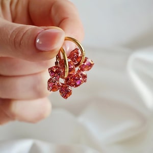 K&ouml;nnte beinhalten: Ein Paar goldfarbene Creolen mit h&auml;ngenden, facettierten Anh&auml;ngern aus durchscheinendem rotem und rosa Harz. Die Anh&auml;nger haben eine glitzernde, funkelnde Textur, die das Licht einf&auml;ngt, und sind an schlichten, polierten goldfarbenen Metallringen befestigt.
