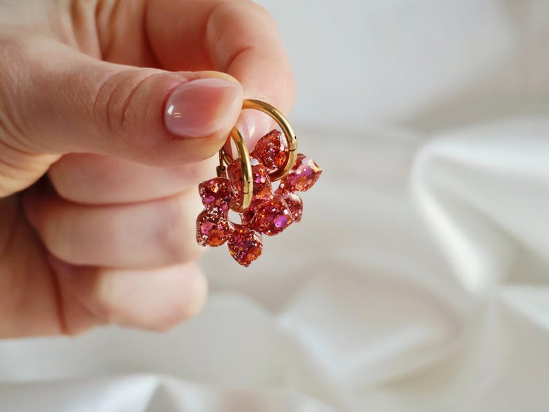 K&ouml;nnte beinhalten: Ein Paar goldfarbene Creolen mit h&auml;ngenden, facettierten Anh&auml;ngern aus durchscheinendem rotem und rosa Harz. Die Anh&auml;nger haben eine glitzernde, funkelnde Textur, die das Licht einf&auml;ngt, und sind an schlichten, polierten goldfarbenen Metallringen befestigt.