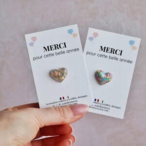 May include: Two white cards with the text "MERCI pour cette belle ann&eacute;e" and a heart-shaped pin. The pins have a glittery, iridescent finish. Pastel balloons are in the upper corners. The cards are held by a hand.