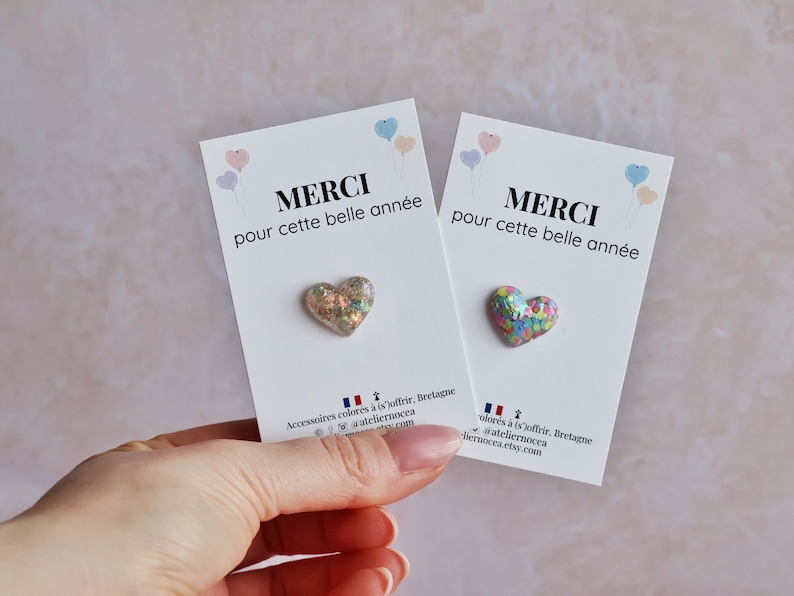 May include: Two white cards with the text "MERCI pour cette belle ann&eacute;e" and a heart-shaped pin. The pins have a glittery, iridescent finish. Pastel balloons are in the upper corners. The cards are held by a hand.