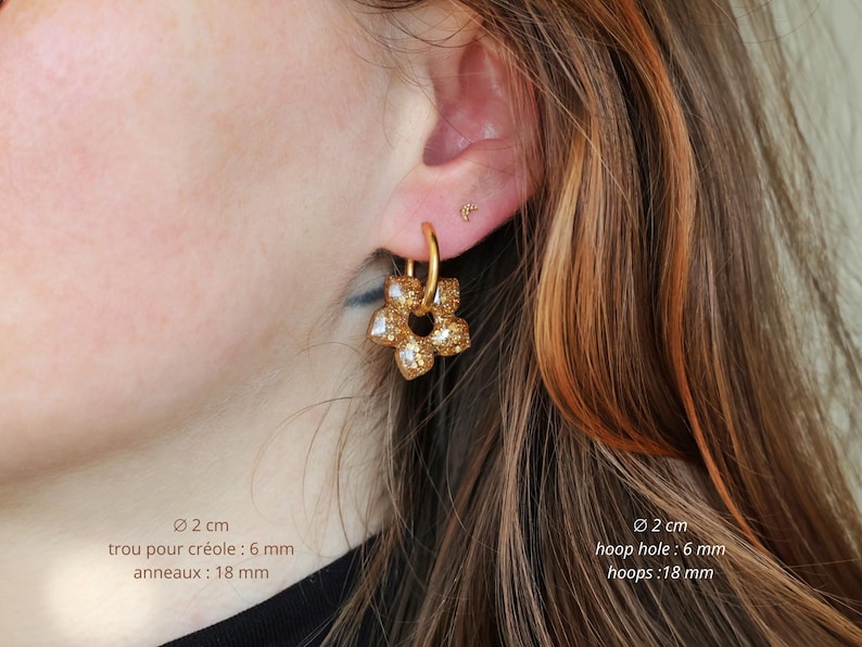 K&ouml;nnte beinhalten: Ein goldfarbener Creolen-Ohrring mit einem f&uuml;nfbl&auml;ttrigen Blumenanh&auml;nger in glitzernder, texturierter Optik. Der Ohrring hat einen Durchmesser von 2 cm, ein 6 mm gro&szlig;es Loch und 18 mm gro&szlig;e Ringe, wobei der Text diese Ma&szlig;e deutlich angibt.