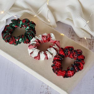 May include: Three fabric scrunchies in festive holiday patterns. One is green with red accents, another is white with red stripes, and the third is red plaid with green and black accents. They are arranged on a neutral-colored surface.