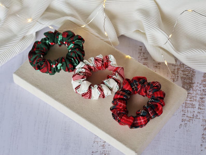 May include: Three fabric scrunchies in festive holiday patterns. One is green with red accents, another is white with red stripes, and the third is red plaid with green and black accents. They are arranged on a neutral-colored surface.