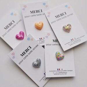 May include: Glitter heart-shaped pins in various colors, including pink, gold, silver, and multicolored, are displayed on white cards. Each card has the French word "MERCI" and the phrase "pour cette belle ann&eacute;e" printed on it.