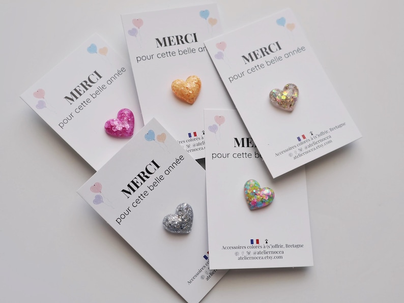 May include: Glitter heart-shaped pins in various colors, including pink, gold, silver, and multicolored, are displayed on white cards. Each card has the French word "MERCI" and the phrase "pour cette belle ann&eacute;e" printed on it.