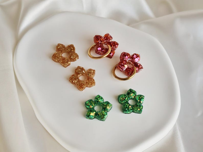 K&ouml;nnte beinhalten: Eine Sammlung von sechs blumenf&ouml;rmigen Anh&auml;ngern aus glitzerndem Harz in Gold, Rot und Gr&uuml;n. Zwei der roten Blumenanh&auml;nger sind an runden, goldfarbenen Metallringen befestigt, w&auml;hrend die restlichen vier Anh&auml;nger einzelne St&uuml;cke ohne Ringe sind.
