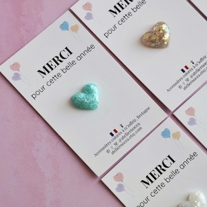 May include: Close-up of several white cards with heart-shaped embellishments. Each card has the French word "MERCI" and the phrase "pour cette belle ann&eacute;e" printed on it. The hearts are various colors and textures.