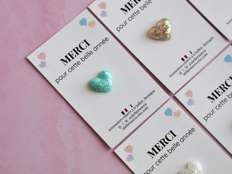 May include: Close-up of several white cards with heart-shaped embellishments. Each card has the French word "MERCI" and the phrase "pour cette belle ann&eacute;e" printed on it. The hearts are various colors and textures.