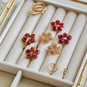 K&ouml;nnte beinhalten: Eine Sammlung von Schmuckst&uuml;cken, bestehend aus vier blumenf&ouml;rmigen Anh&auml;ngern mit rotem und goldenem Glitzer, zwei goldfarbenen Creolen, einem schlichten Goldring und einem Goldring mit drei kleinen klaren Steinen, angeordnet in einer Schmuckschatulle.
