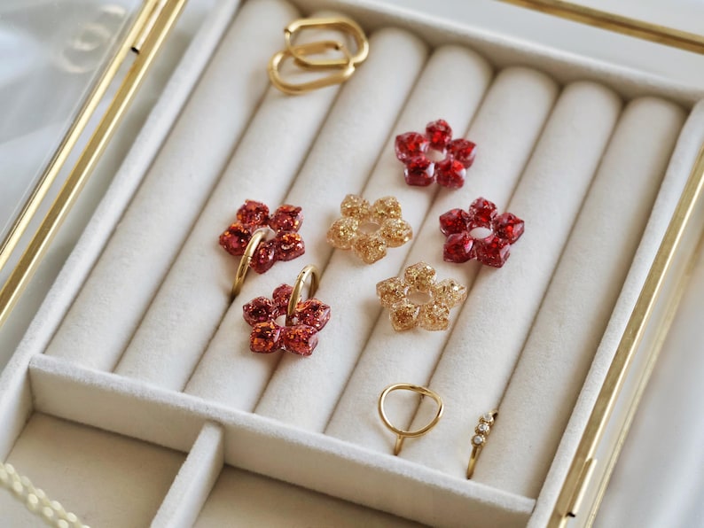K&ouml;nnte beinhalten: Eine Sammlung von Schmuckst&uuml;cken, bestehend aus vier blumenf&ouml;rmigen Anh&auml;ngern mit rotem und goldenem Glitzer, zwei goldfarbenen Creolen, einem schlichten Goldring und einem Goldring mit drei kleinen klaren Steinen, angeordnet in einer Schmuckschatulle.