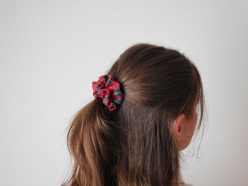 May include: A red, green, and white plaid scrunchie is tied in a low ponytail. The hair accessory is made of a soft fabric and adds a pop of color to the hairstyle. The scrunchie is a classic hair accessory.